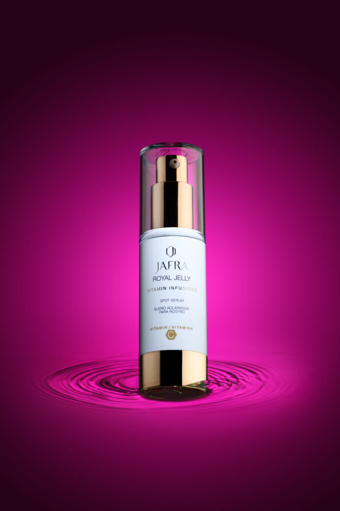 Flacon de produit de beauté de marque Jafra disposé sur un fond dégradé de rose fuschia au noir à l'extérieur, en légére contre plongée, le socle du flacon est entouré d'un mouvement d'eau circulaire